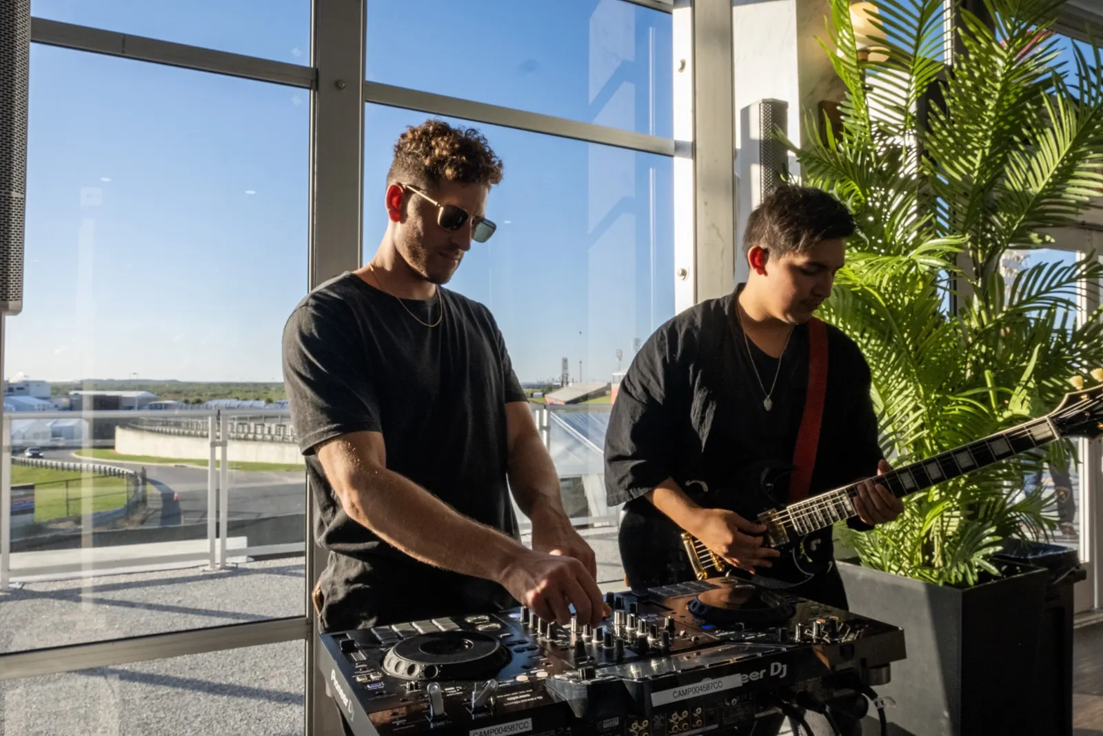 Jordan Miller on the decks beside a live electric guitarist in a glass-walled suite overlooking a racetrack.