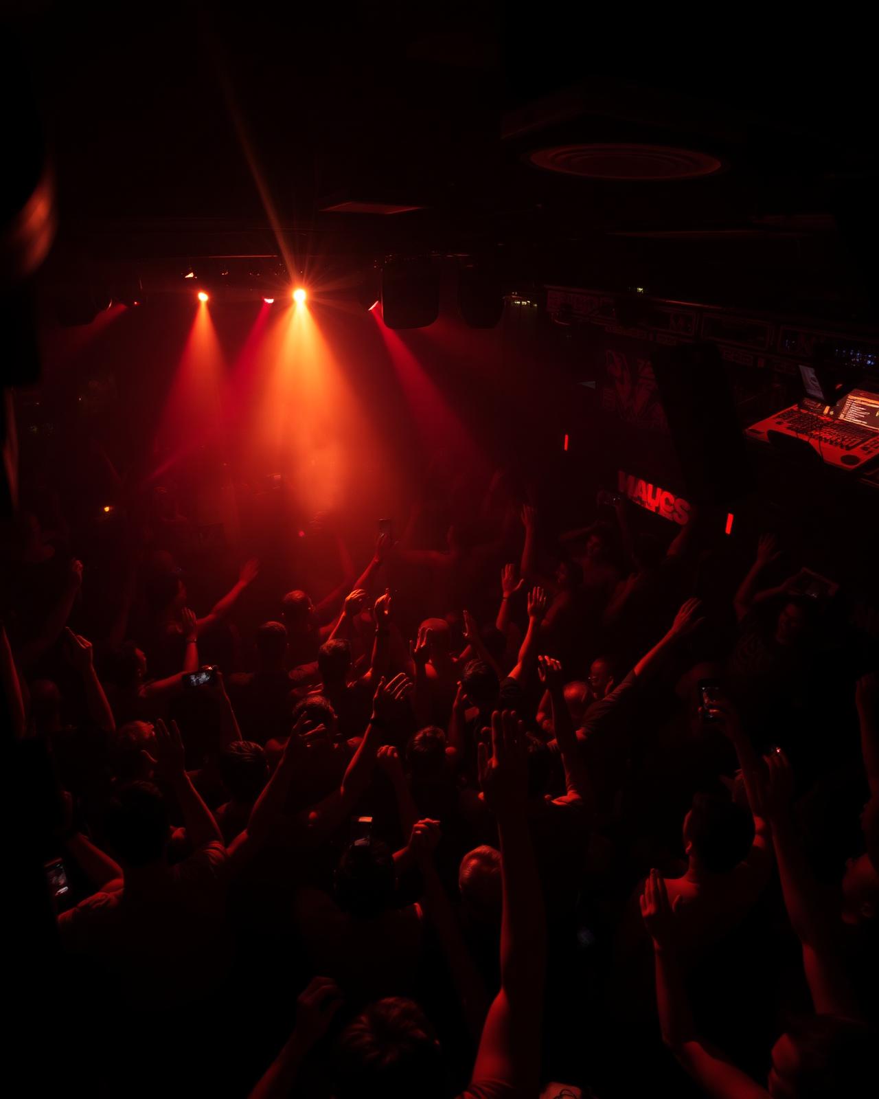 A packed club dance floor with hands up under deep red stage beams, the DJ booth glowing at the right.